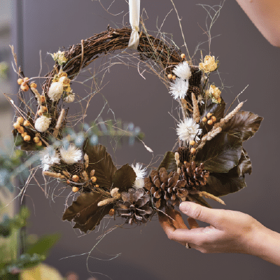 Nov 10, Natural Materials Workshop: Hand holding a dried wreath of twigs, pinecones, and dried flowers.